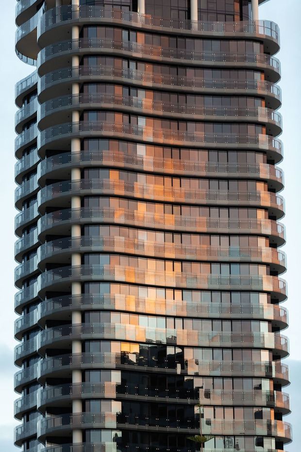 Close-up exterior view of a tall contemporary apartment building with a rounded tower form, extensive floor-to-ceiling glazing, continuous curved balconies, transparent glass balustrades, and dark grey structural bands. Warm reflections from the surrounding city or sunset appear across the windows.