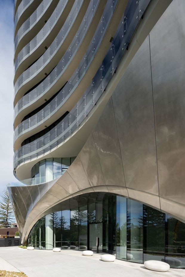 Exterior architectural photograph of a contemporary multi-storey building with dramatically curved, stacked balconies enclosed by clear glass balustrades. The lower facade features sculptural sweeping curves, large floor-to-ceiling glazed entry walls, and metallic champagne-toned cladding. A paved forecourt with low sculptural seating is visible at ground level.