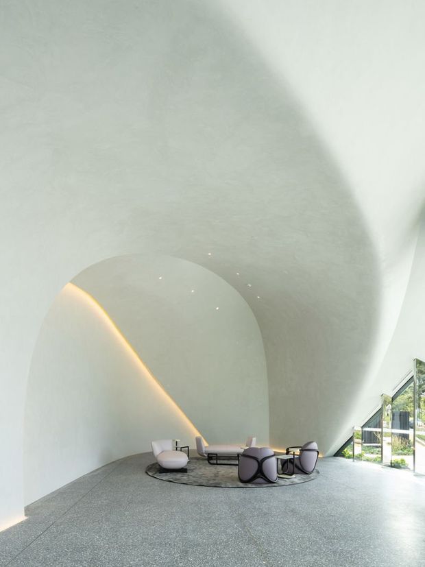 A sculptural, highly curved interior lounge with seamless pale plaster walls and ceiling forming an organic vaulted shell. A warm concealed LED strip traces the sweeping curve, while a low circular coffee table, contemporary sculptural lounge chairs, and a neutral rug create a small seating area. The speckled grey floor and glazed opening to the landscaped exterior reinforce the bright, minimalist gallery-like atmosphere.