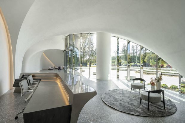 A refined modern reception or lounge interior with a sculptural curved white ceiling and walls, expansive floor-to-ceiling glazing, a long built-in dark reception counter with integrated warm lighting, and a circular seating area on a grey rug. The space features polished terrazzo-like flooring, a central cylindrical column, minimalist lounge chairs, and views to landscaped grounds outside.