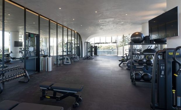 A spacious, modern gym with dark rubber flooring, extensive strength-training and free-weight equipment, full-height glass partitions and glazing, and a textured concrete ceiling. The open layout is brightly lit by recessed ceiling lights and natural daylight, with views toward landscaped surroundings beyond the glass.