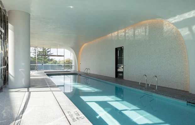 An indoor swimming pool in a bright, sculptural modern space. The long rectangular pool is bordered by pale terrazzo-like paving, with stainless-steel handrails and a textured curved wall enclosing one side. Large glazed openings bring in strong daylight and views of vegetation outside, while a rounded column and curved ceiling reinforce the organic modern architecture.