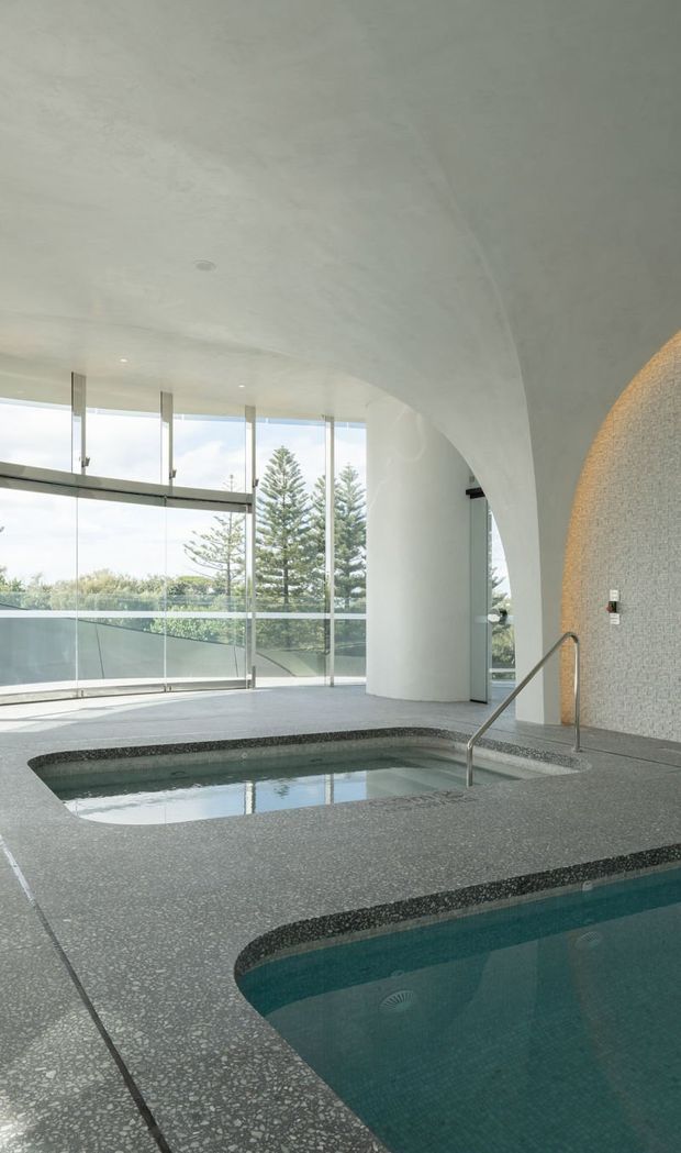 An airy indoor pool and spa area with curved white architectural forms, full-height glazing, and views of trees outside. The pools are finished with grey terrazzo-like aggregate surfaces and dark contrasting edge bands, with a stainless-steel handrail leading into the water. The space has a calm, refined, resort-like atmosphere and abundant natural light.