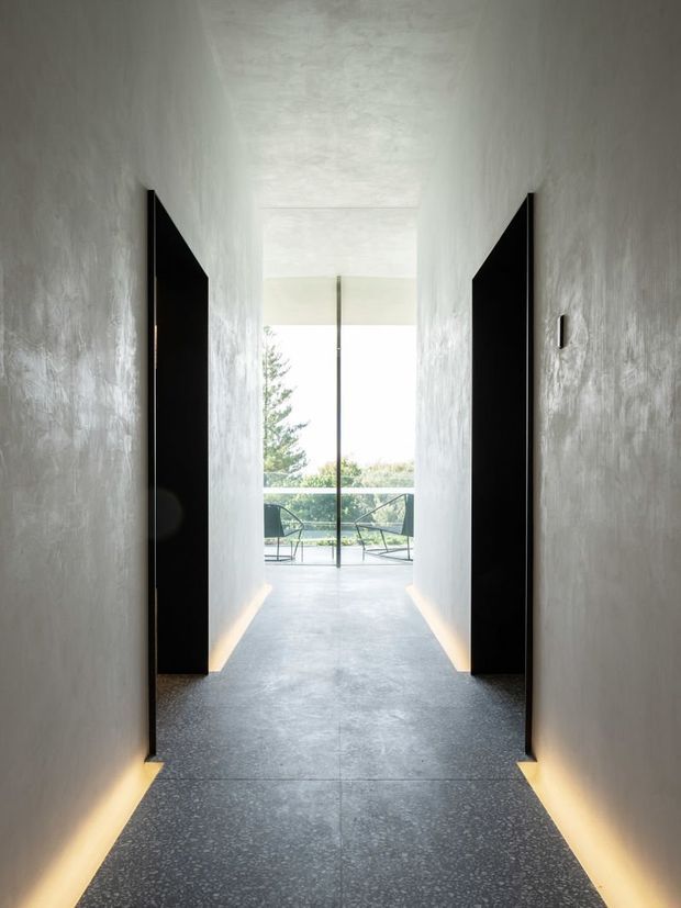 Minimalist hallway with textured light-grey plaster walls, dark flush doors, concealed warm floor-level lighting, and a polished speckled grey floor. The corridor leads to floor-to-ceiling glazing and a balcony or terrace with outdoor seating.