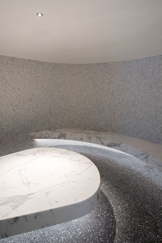 A curved, intimate spa-like interior featuring small grey mosaic-tiled walls and flooring, a large rounded white marble-clad platform or bench, and a curved marble ledge around the perimeter. A recessed ceiling spotlight provides subdued illumination. The space has a restrained, monochromatic and highly tactile material palette.