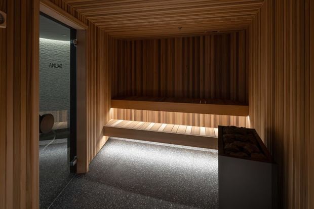 Compact sauna interior clad in warm vertical timber slats, with a timber bench, softly concealed lighting beneath the seating, and a dark textured floor. A glass door opens to an adjacent wet area, visible at the left.