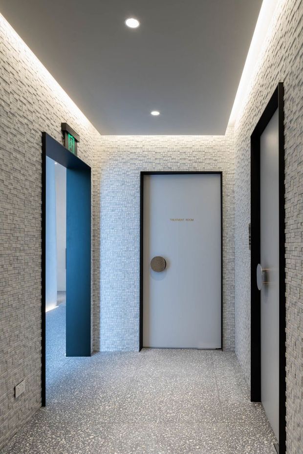 A narrow contemporary interior corridor with white textured masonry or small-format tile wall finishes, a speckled terrazzo-like floor, dark-framed doors, and concealed linear cove lighting around a dark ceiling. A blue-lined doorway with an illuminated exit sign opens on the left, while a central frosted door is labelled “Treatment Room.”