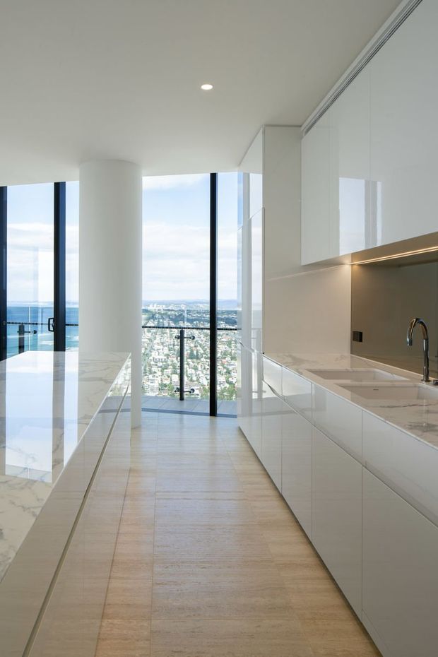 A bright, minimalist contemporary kitchen in a high-rise apartment, featuring a long white stone island, white handleless cabinetry, integrated under-cabinet lighting, a pale timber-look floor, and full-height glazing with expansive city and water views. A curved structural column and balcony are visible beyond the kitchen.