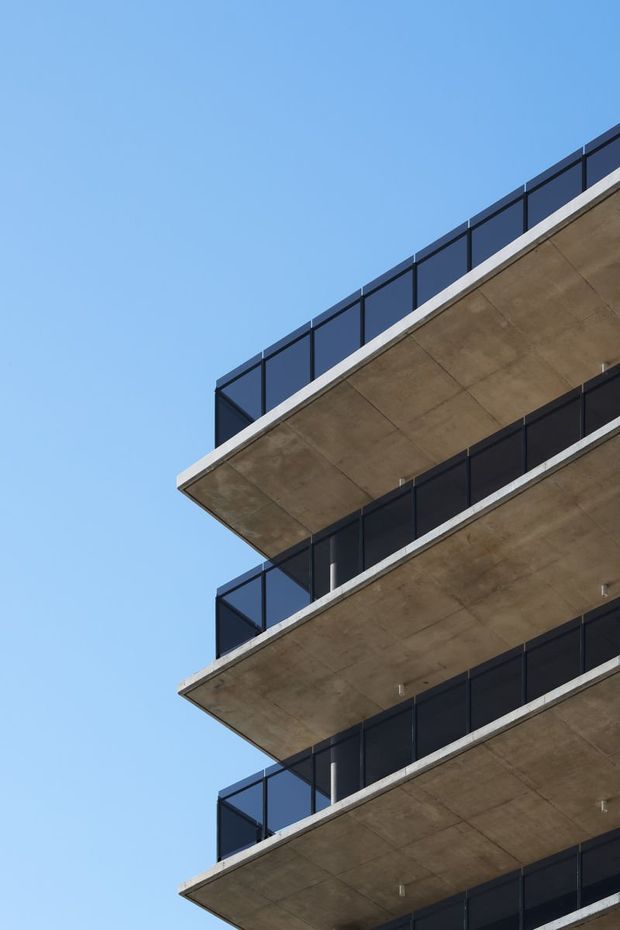 Upward-looking close-up of a modern multi-storey building with stacked projecting concrete balconies and dark framed glass or solid-panel balustrades against a clear blue sky. The image emphasizes the repetitive cantilevered balcony structure and minimalist facade.