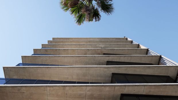Upward-looking view of a contemporary multi-storey concrete building, showing stacked projecting balcony slabs, dark glazing, glass balustrades, and a palm frond against a clear blue sky.