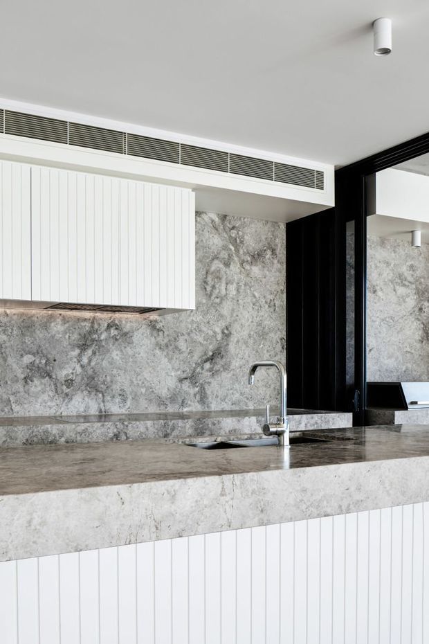 Close-up view of a contemporary kitchen featuring a large grey stone island benchtop with integrated sink and chrome tap, matching stone splashback and wall surfaces, white vertically fluted cabinetry, black-framed glazing or joinery, and a minimalist white ceiling with linear air-conditioning grilles and recessed spotlight.