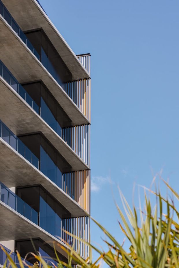 Upward-looking exterior view of a contemporary multi-storey building with projecting concrete balcony slabs, dark glazed balcony fronts, and vertical metal screening elements. A clear blue sky and foreground planting frame the facade.