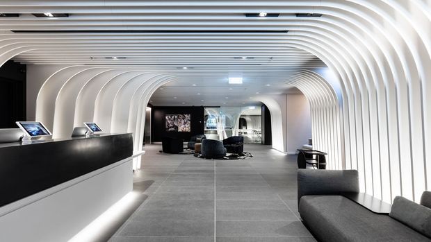 A sleek contemporary commercial interior featuring a reception counter, lounge seating, and a dramatic tunnel-like corridor formed by continuous white curved ribbed wall and ceiling panels. Dark grey tiled flooring, black reception joinery, integrated linear lighting, and a bright monochromatic palette create a refined, futuristic hospitality or showroom environment.