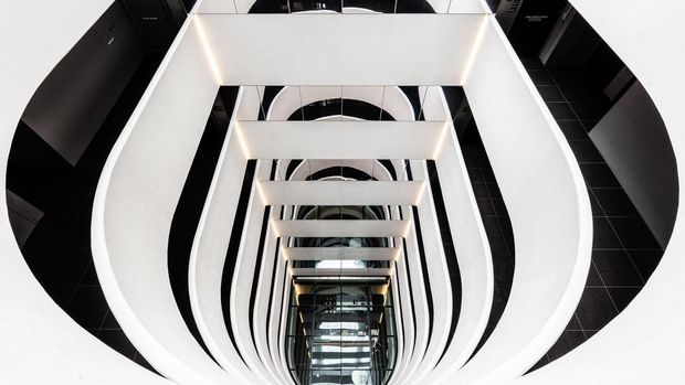 Looking upward through a dramatic multi-level interior atrium or stairwell, with repeating white curved structural balustrades and rectangular floor openings creating a strong geometric perspective. Glossy black tiled surfaces, mirrored reflections, and linear integrated lighting produce a bold, futuristic architectural composition.