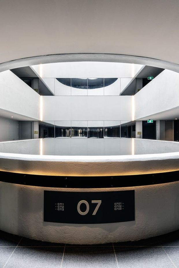 A symmetrical, modern multi-level interior atrium or hallway with curved white walls, dark glazed balustrades, integrated linear lighting, and a prominent black level-07 directory sign. The space has a polished commercial or institutional character, with visible lift or corridor doors and exit signage.