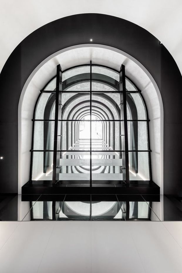 A dramatic upward view through a tall, arched architectural atrium or stairwell. Repeated curved glass arches, black structural framing, reflective glazing and white walls create a strong geometric perspective and bright central vanishing point. The space is monochromatic, highly symmetrical and appears to have significant vertical scale.