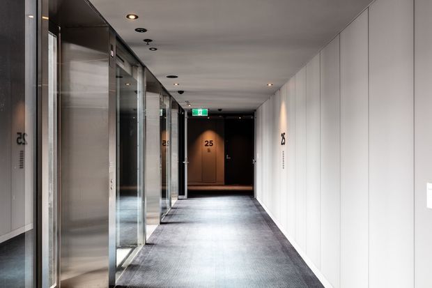 A modern commercial hallway with a long, narrow corridor, dark carpet flooring, white smooth wall panels, recessed ceiling lights, and glazed lift or office fronts along the left side. Dark timber-toned doors and room numbers are visible at the far end, with bright linear perspective and understated contemporary detailing.