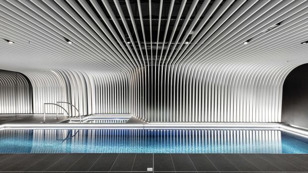 A dramatic indoor swimming pool enclosed by a continuous white ribbed architectural ceiling and wall treatment. The curved vertical fins wrap around the pool enclosure and extend overhead, creating a sculptural tunnel-like form. The pool has calm blue water, integrated linear lighting, and a dark tiled surround.