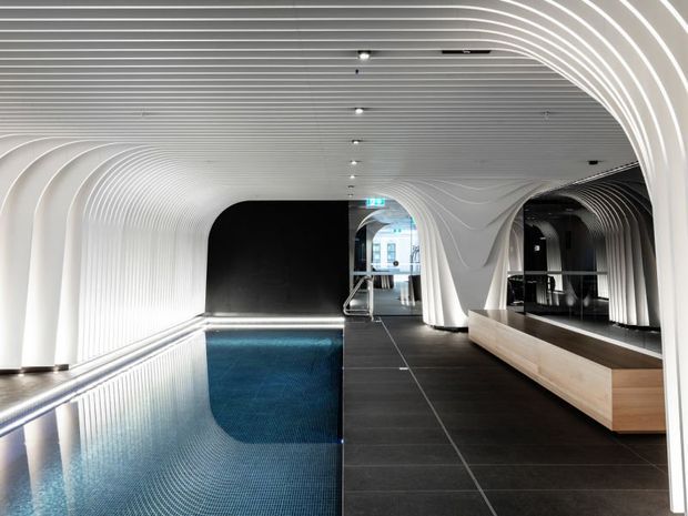 A striking contemporary interior featuring a narrow indoor swimming pool beside a dark tiled passage. Sculptural white ribbed wall and ceiling panels form flowing curved arches and tunnel-like transitions, with integrated linear lighting and a light timber reception or bench element. The space appears to be a high-end wellness, spa, or hospitality facility.