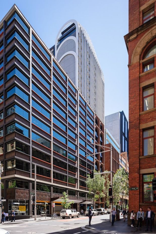 Street-level view of a dense urban commercial district with several mid- and high-rise buildings. The prominent contemporary buildings feature glazed curtain walls, dark horizontal bands, red-brown masonry accents, and a distinctive rounded white tower crown. A historic red-brick building with arched window detailing frames the right side, while pedestrians, street trees, traffic signals, and storefronts occupy the foreground.
