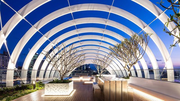 A contemporary rooftop terrace or elevated outdoor walkway beneath a dramatic series of curved white structural arches with blue translucent or open panels. The space features timber decking, illuminated built-in planters with small trees, integrated bench seating, metal balustrades, and a city skyline beyond. The scene is bright, refined, and strongly architectural, with a tunnel-like vaulted canopy creating a rhythmic perspective.