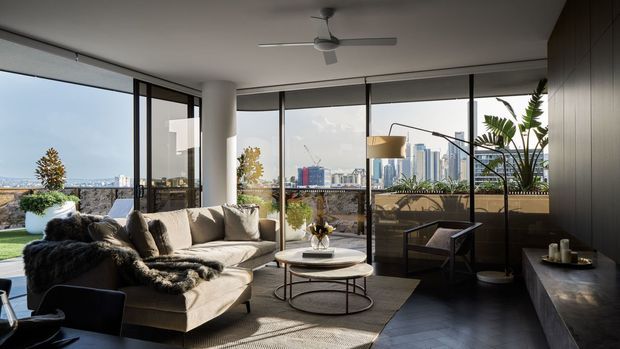 A sophisticated contemporary living room with expansive floor-to-ceiling glazing overlooking a dense urban skyline. The room features a large neutral sectional sofa, layered textured throws and cushions, a round coffee table, a sculptural floor lamp, dark built-in wall cabinetry, a ceiling fan, and a balcony with greenery visible beyond the glass. The space is bright, refined, and strongly connected to the city view.