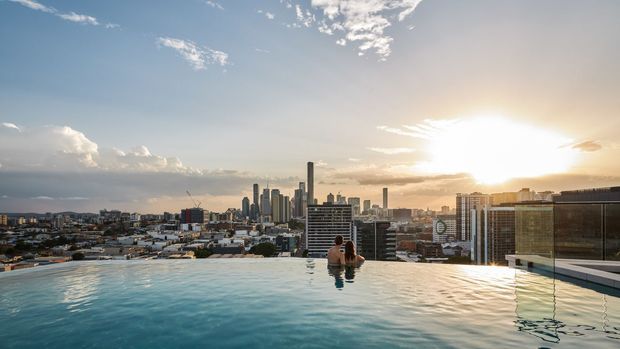 A rooftop infinity pool overlooking a dense modern city skyline at sunset. Two people are partially visible in the pool, while high-rise residential and commercial buildings extend across the urban horizon. The scene has warm golden sunlight, reflective water, and expansive metropolitan views.
