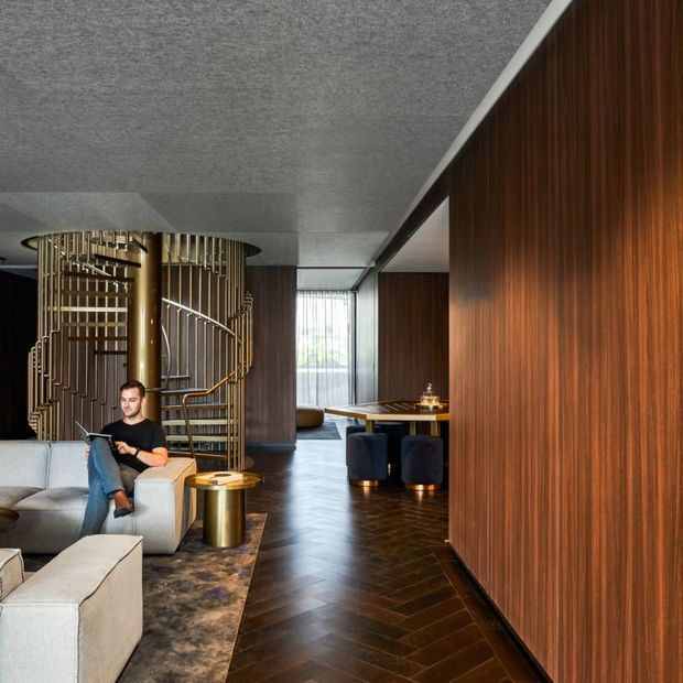 A sophisticated open-plan interior combining a lounge and dining area. The space features dark timber wall panelling and herringbone flooring, a sculptural brass-toned spiral staircase with decorative metal balustrade, a pale sectional sofa, a metallic side table, and a compact dining setting. Full-height glazing at the rear brings in natural light, while the overall palette is dark, warm, and refined.
