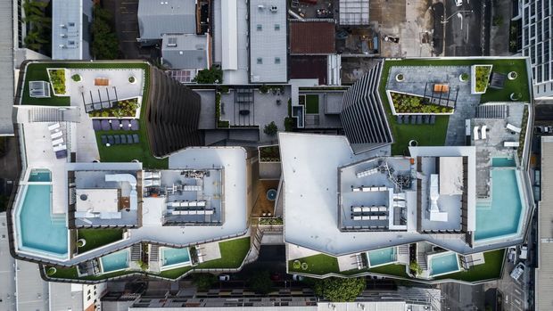 Aerial top-down view of a dense urban multi-storey building complex with several rooftop terraces, landscaped green roofs, outdoor seating areas, rooftop equipment, and multiple turquoise swimming pools. The contemporary flat-roofed structures are tightly arranged within a city block.