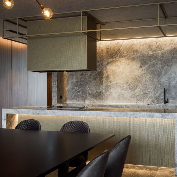 Moody, refined open-plan kitchen and dining space with a dark dining table and upholstered chairs in the foreground. The kitchen features a long stone island and matching stone splashback, textured metallic cabinetry or screening, dark timber wall panelling, black tapware, and an exposed dark metal ceiling framework with warm pendant globes.