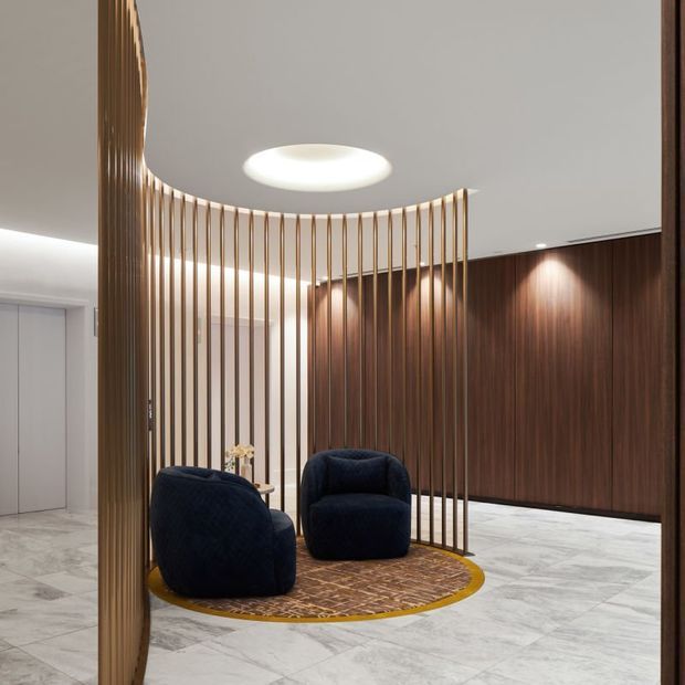A refined lobby or reception area featuring a curved screen of slender vertical timber slats, dark timber wall panelling with integrated lighting, pale marble-look flooring, and two deep navy lounge chairs arranged on a circular patterned rug with a brass-gold border. A sculptural recessed ceiling light adds a soft focal point.