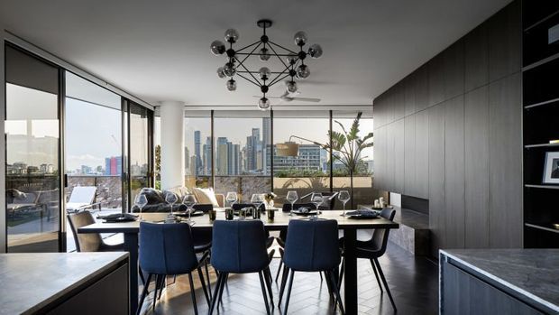 Contemporary open-plan dining area with a long dining table and deep blue upholstered chairs, overlooking a dense city skyline through expansive floor-to-ceiling glazing. Dark built-in cabinetry lines the right wall, with a stone-topped island or counter in the foreground and a sculptural multi-arm pendant light overhead. A balcony or terrace with plants is visible beyond the glazing.
