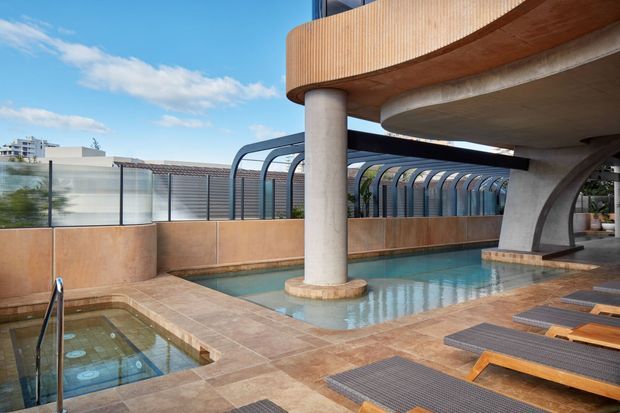 A rooftop or elevated pool area with a main swimming pool, a smaller spa pool, tiled surrounds, timber sun loungers, curved concrete columns, and a distinctive arched glazed or louvred enclosure. Warm terracotta and timber finishes contrast with the blue water and sky.