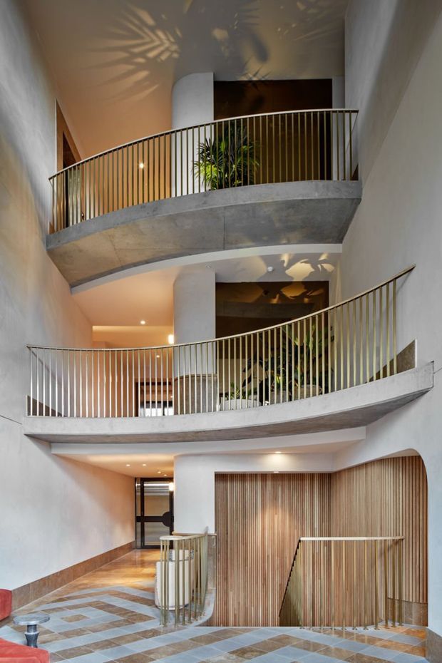 A multi-level interior atrium or stair hall with sweeping curved concrete balconies, slim vertical metal balustrades, warm timber slat wall cladding, white plaster walls, and patterned tile flooring. Several planted areas and dramatic concealed lighting create a sculptural, contemporary circulation space.