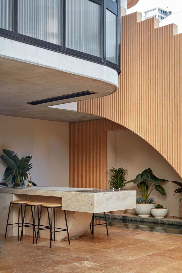 A refined modern interior featuring a sculptural kitchen island with a pale stone benchtop, integrated sink and brass tap, and black bar stools. Warm terracotta-toned stone flooring, ribbed timber wall cladding, curved concrete forms, dark-framed glazing, and abundant indoor plants create a warm contemporary atmosphere. A staircase with a curved ribbed timber underside is visible at right.