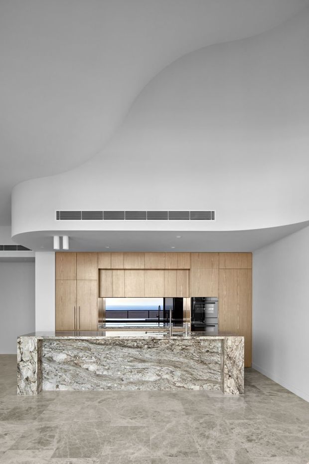 A refined contemporary kitchen with a large natural-stone island, full-height light timber cabinetry, integrated appliances, and a sculptural curved white ceiling bulkhead. The space has polished stone flooring, discreet linear air-conditioning vents, and a restrained neutral palette.