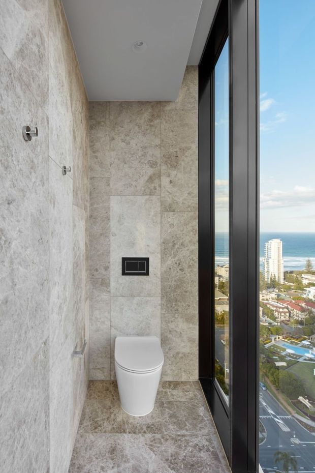A narrow, contemporary toilet room with full-height light stone-look wall and floor tiles, a wall-hung white toilet, concealed flush plate, and a tall black-framed window offering an expansive coastal city and ocean view. The space is clean, minimal, and refined, with recessed ceiling lighting and brushed metal wall fittings.
