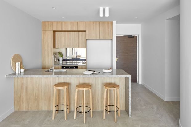 Bright, compact contemporary kitchen with a light timber island featuring three stools, a pale stone benchtop, integrated timber cabinetry, stainless-steel sink and tap, and a dark timber entry door beyond. Light grey walls and flooring create a clean, neutral scheme.
