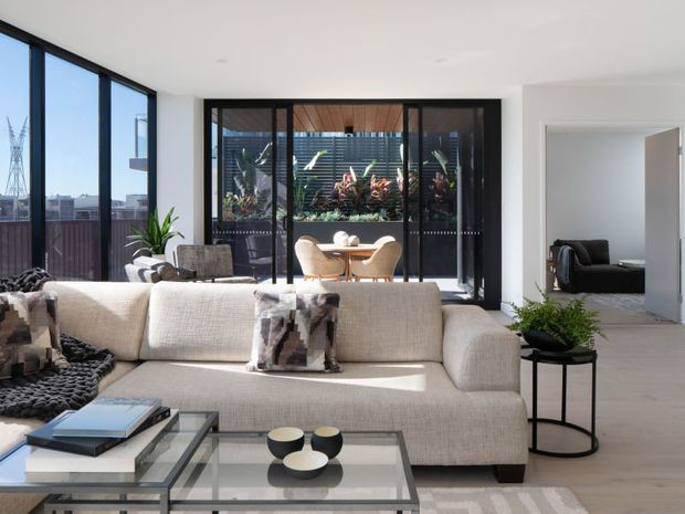 Bright contemporary open-plan living room with a large cream sectional sofa, glass coffee tables, neutral styling, and floor-to-ceiling black-framed glazing. A dining area and planted courtyard are visible beyond the internal glass partition, with a bedroom or secondary room visible through the doorway on the right.