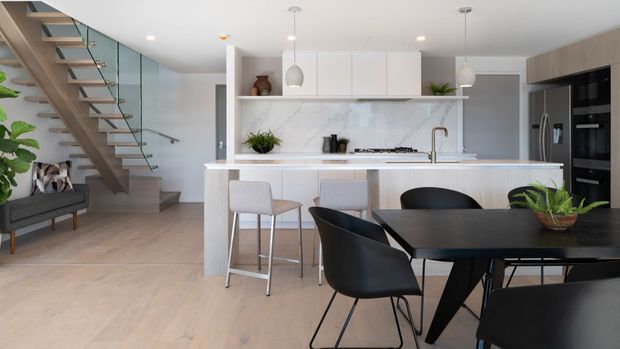 A bright, contemporary open-plan kitchen and dining area with a white island and marble-look splashback, integrated dark cabinetry, pale timber flooring, and a glass-balustraded timber staircase. A black dining table with modern chairs occupies the foreground, while pendant lights and indoor plants add subtle accents.