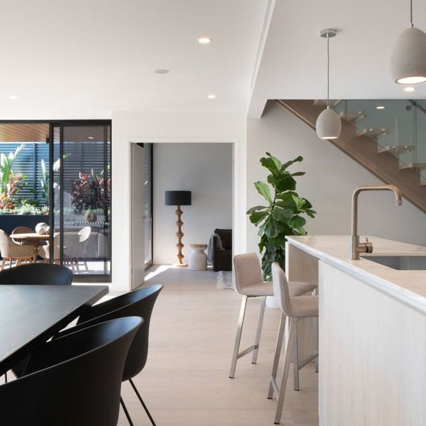 Bright open-plan contemporary interior showing a kitchen island with pale cabinetry and stone benchtop, adjacent dining area with a dark table and black chairs, and a glazed outdoor sitting area beyond. A stairway with timber detailing, pendant lights, indoor plants, and a separate living nook are also visible.