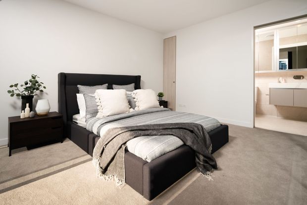 A bright, contemporary bedroom with a large upholstered black bed and matching footstool, layered grey and white bedding, dark bedside furniture, and neutral carpet. A simple white door and an open ensuite bathroom with a floating vanity are visible on the right.