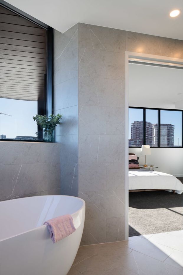 A bright contemporary bathroom with a freestanding white bathtub, large pale stone-look wall and floor tiles, a black-framed window, and recessed ceiling lighting. The bathroom opens directly into a bedroom visible through the doorway, with floor-to-ceiling glazing and city views.