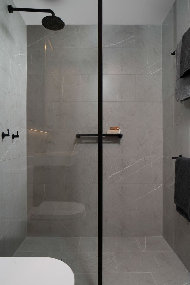 A compact contemporary bathroom dominated by a walk-in shower enclosed with a clear glass screen and black metal framing. The walls and floor are clad in large-format pale grey marble-look tiles with subtle veining. Matte black shower fittings, towel hooks, a shelf, and dark grey towels provide contrast; the edge of a white freestanding bathtub is visible in the foreground.