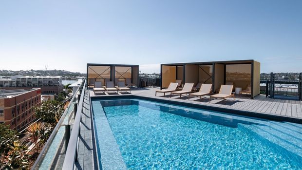 A rooftop swimming pool on a contemporary multi-storey building, surrounded by a timber-toned deck, sun loungers, and dark-framed poolside cabana structures. The elevated terrace overlooks a dense urban neighbourhood and waterfront-like city surroundings under a clear blue sky.