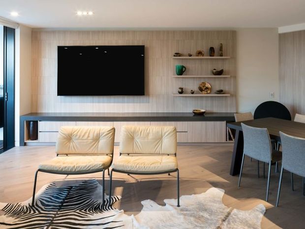 Contemporary open-plan living and dining area with a wall-mounted television set into a light timber feature wall, integrated low cabinetry, open display shelving, and a long dark benchtop-like console. Two cream lounge chairs sit over layered animal-print rugs, while a dark dining table with upholstered chairs occupies the right side. Large glazing at the left brings in abundant natural light.