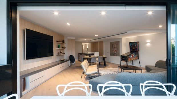 Open-plan contemporary living and dining area viewed through large dark-framed glazing. The room features pale timber wall panelling and flooring, a built-in media wall with television and low cabinetry, a neutral sofa and lounge chairs, a dining table, recessed ceiling lighting, and contemporary artwork and furnishings.