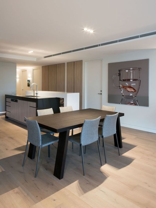 A bright, contemporary open-plan dining and kitchen area. A large dark timber dining table with grey upholstered chairs sits on a pale timber floor. Behind it is a streamlined kitchen with a dark island, white benchtop, tall timber-toned cabinetry, integrated appliances, and minimalist white walls. Recessed ceiling lighting, linear air-conditioning vents, and a large abstract artwork complete the refined interior.