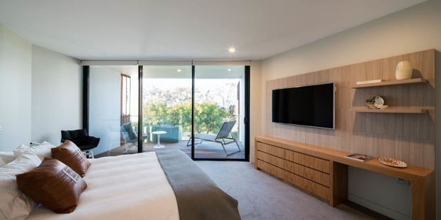 A contemporary bedroom with a large upholstered bed, white bedding and brown cushions, facing a full-width timber media wall with built-in cabinetry, open shelving and a wall-mounted television. Floor-to-ceiling glazed doors open to a balcony or terrace, bringing in abundant natural light. The room has soft carpet flooring, pale walls and restrained modern furnishings.