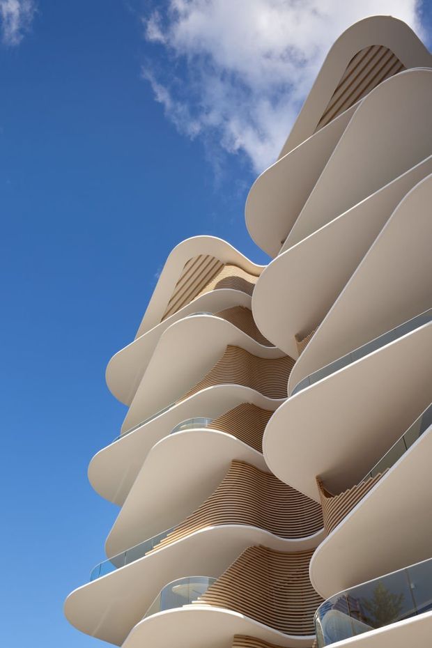 Low-angle architectural photograph of a tall contemporary multi-storey building with dramatically rounded, flowing cream-coloured balcony slabs, curved glass balustrades, and extensive warm timber-toned slatted soffits. The sculptural facade rises against a vivid blue sky with scattered clouds.